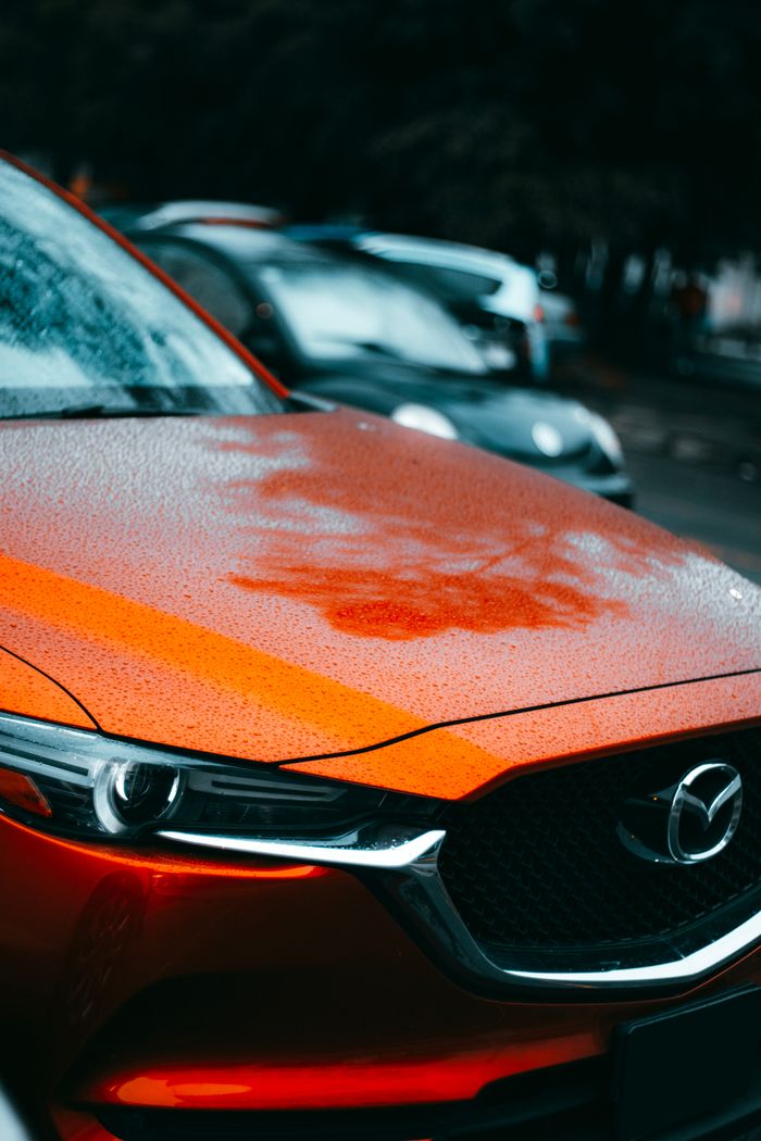 Detailed view of a red car's bonnet with raindrops, showcasing its sleek design and vibrant color in a rainy outdoor setting.